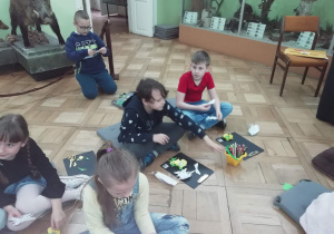 Dzieci uczestniczą w zajęciach warsztatowych siedząc na podłodze i wykonując zadanie